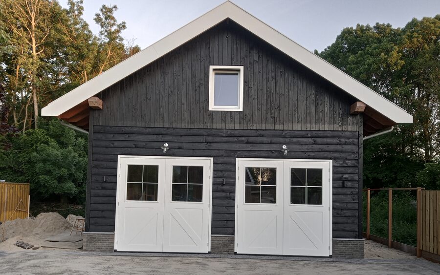 A newly built barn clad in black timber on a brick plinth, with a white bargeboard gable, a dormer window, exposed rafter tails on timber corbels and two pairs of white glazed double doors