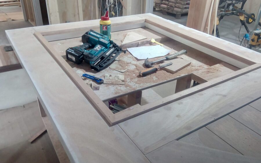 A large frame-and-panel barn door lying flat on trestles in a timber-framed workshop, with a nailer, chisels and glue standing on it