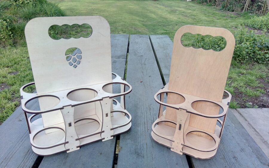 Two flat-pack plywood six-bottle carriers on a garden table, the left one with a hop cone cut through its handle panel