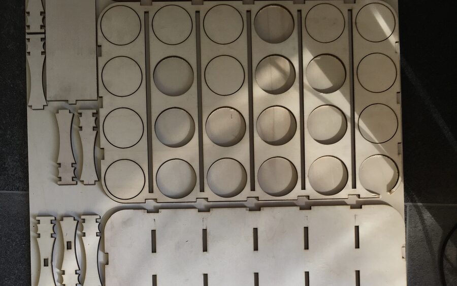A sheet of plywood laser-cut into crate parts — rows of circular bottle openings, slotted panels and curved dividers — all still lying flat in the sheet