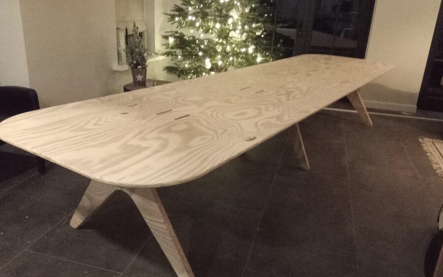 A long plywood dining table with a tapered top standing on splayed cross legs